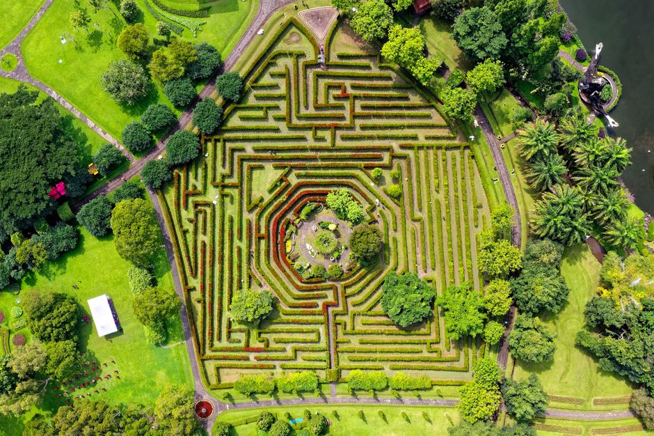Aerial view of a symmetrical formal garden with neatly trimmed boxwood hedges forming intricate maze patterns centered around a circular garden bed with small trees and shrubs. Surrounding the maze ar